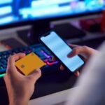 Joven con tarjeta bancaria y móvil frente a un ordenador, representando conducta de riesgo asociada al juego online
