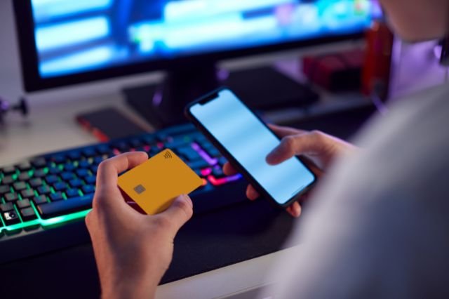 Joven con tarjeta bancaria y móvil frente a un ordenador, representando conducta de riesgo asociada al juego online
