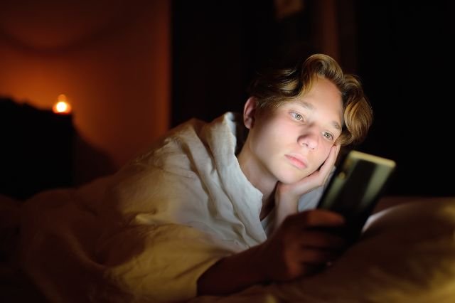 Adolescente acostado mirando su móvil con expresión preocupada, ilustrando el impacto emocional del sexting y grooming